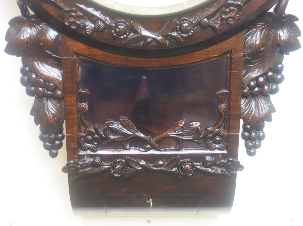 An antique Victorian carved dark mahogany wall clock featuring an off-white dial with Roman numerals marked Berry Cardiff, framed by thistle and vine carvings. View III