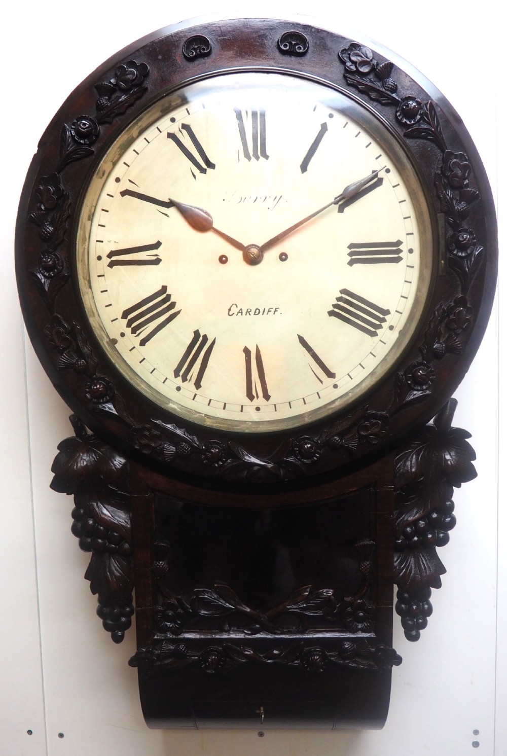 An antique Victorian carved dark mahogany wall clock featuring an off-white dial with Roman numerals marked Berry Cardiff, framed by thistle and vine carvings. View XI