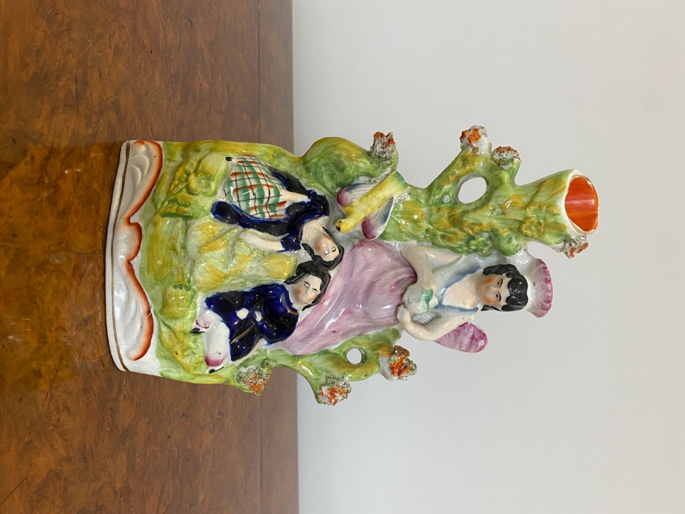 A Victorian Staffordshire earthenware flatback figure featuring three seated figures positioned in front of a tree trunk spill vase with green, pink, and blue polychrome enamel decoration. View II
