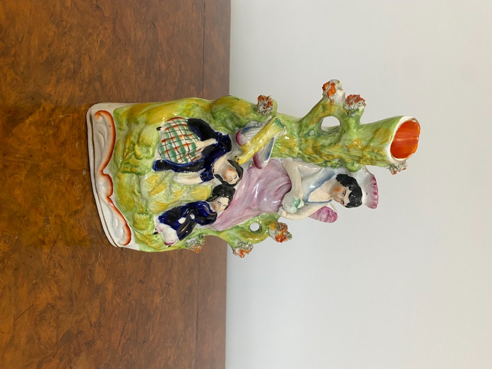 A Victorian Staffordshire earthenware flatback figure featuring three seated figures positioned in front of a tree trunk spill vase with green, pink, and blue polychrome enamel decoration. View III