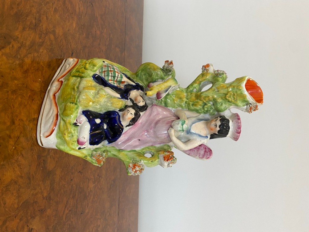 A Victorian Staffordshire earthenware flatback figure featuring three seated figures positioned in front of a tree trunk spill vase with green, pink, and blue polychrome enamel decoration. View IV