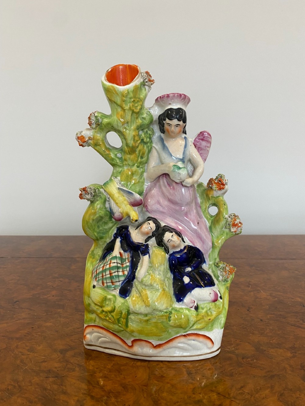 A Victorian Staffordshire earthenware flatback figure featuring three seated figures positioned in front of a tree trunk spill vase with green, pink, and blue polychrome enamel decoration.