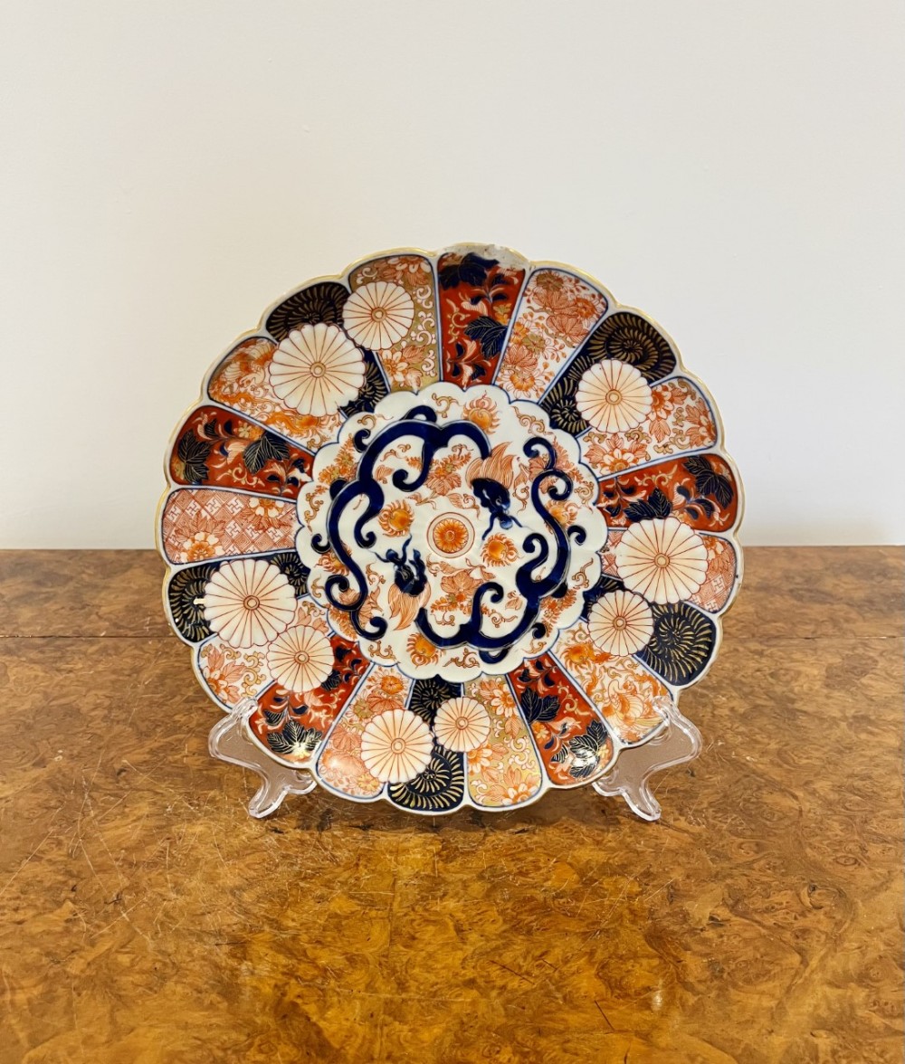 A circular Japanese Imari porcelain plate featuring a scalloped edge, segmented floral panels in terracotta and navy blue, and central gilt embellishments.