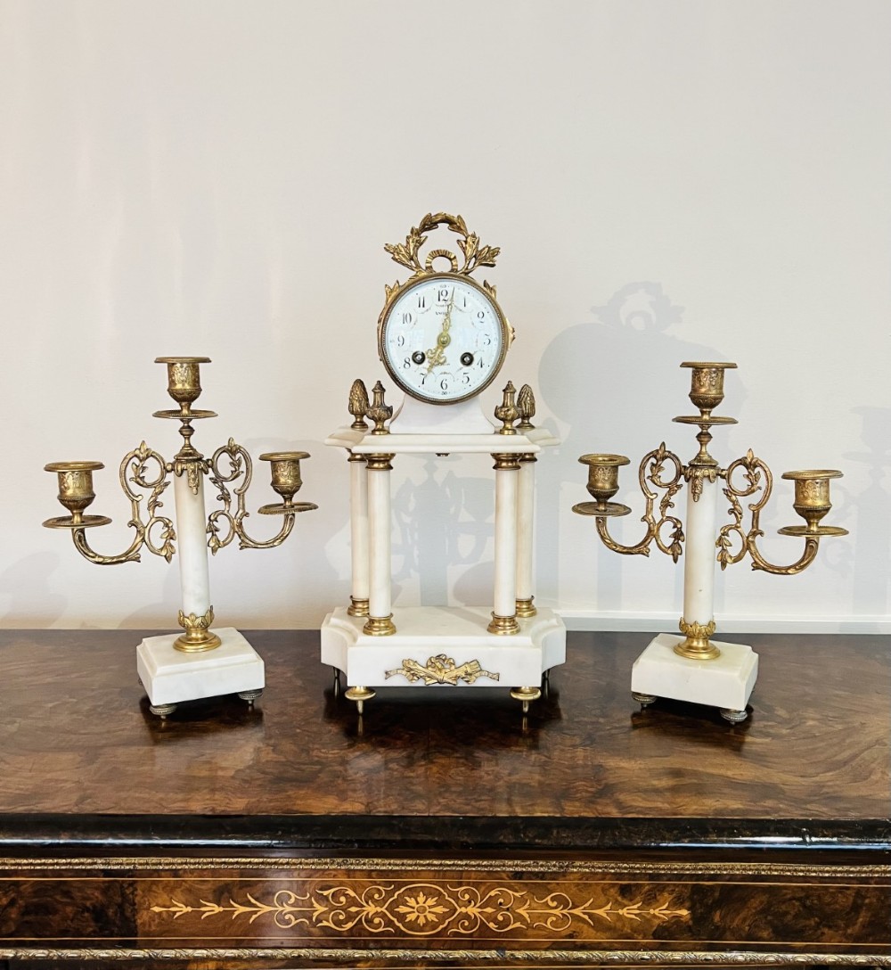 A Victorian three-piece clock garniture crafted from white marble and gilt bronze, featuring a central portico clock supported by four columns alongside two matching three-light candelabras.