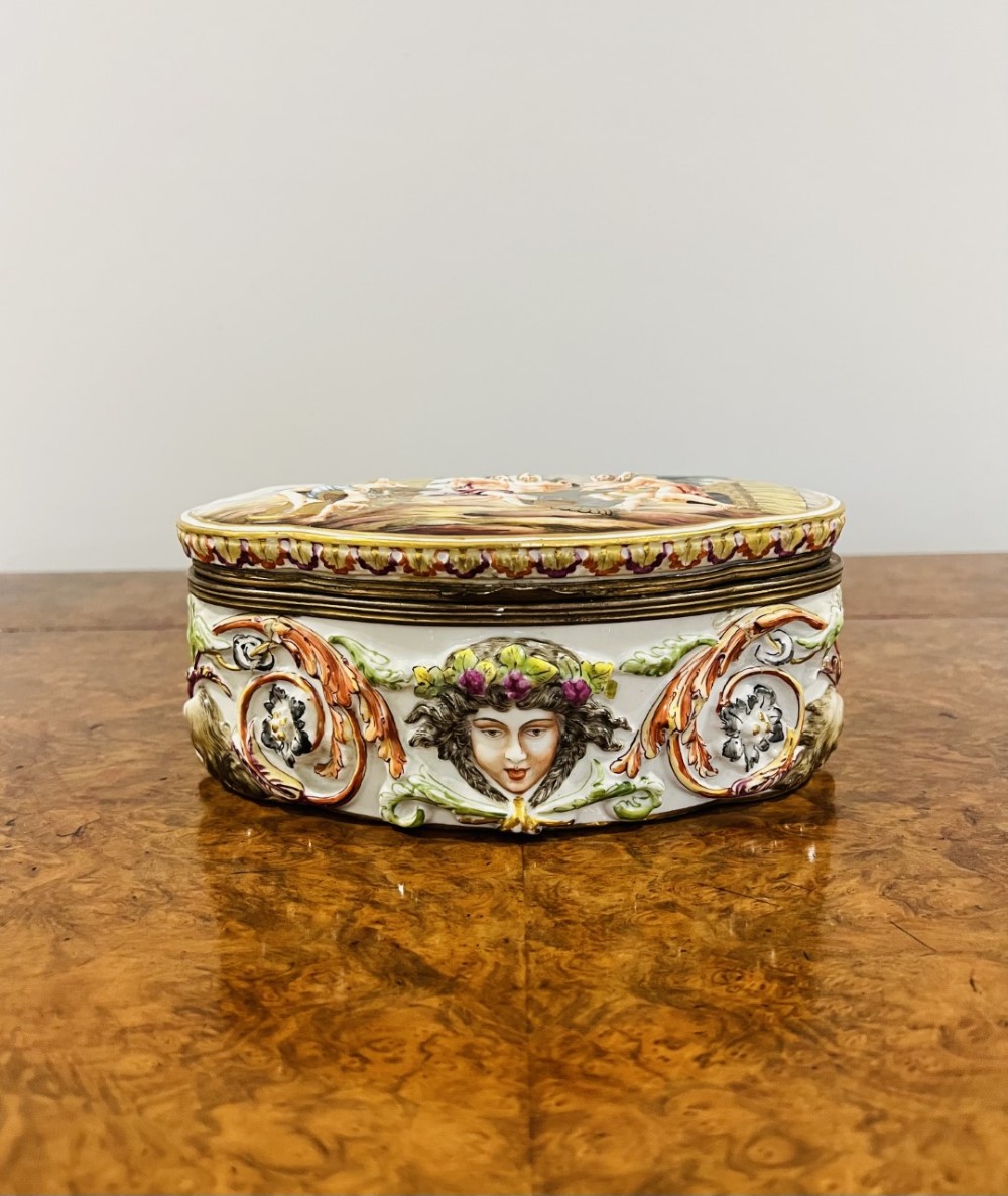 An oval porcelain table casket decorated with hand-painted high-relief classical figures, mascarons, foliage, and fitted with a brass rim and hinge.