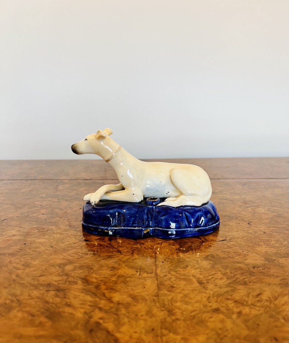 A Victorian Staffordshire glazed earthenware figure depicting a cream reclining greyhound set upon an oval cobalt blue base with a central quill aperture.