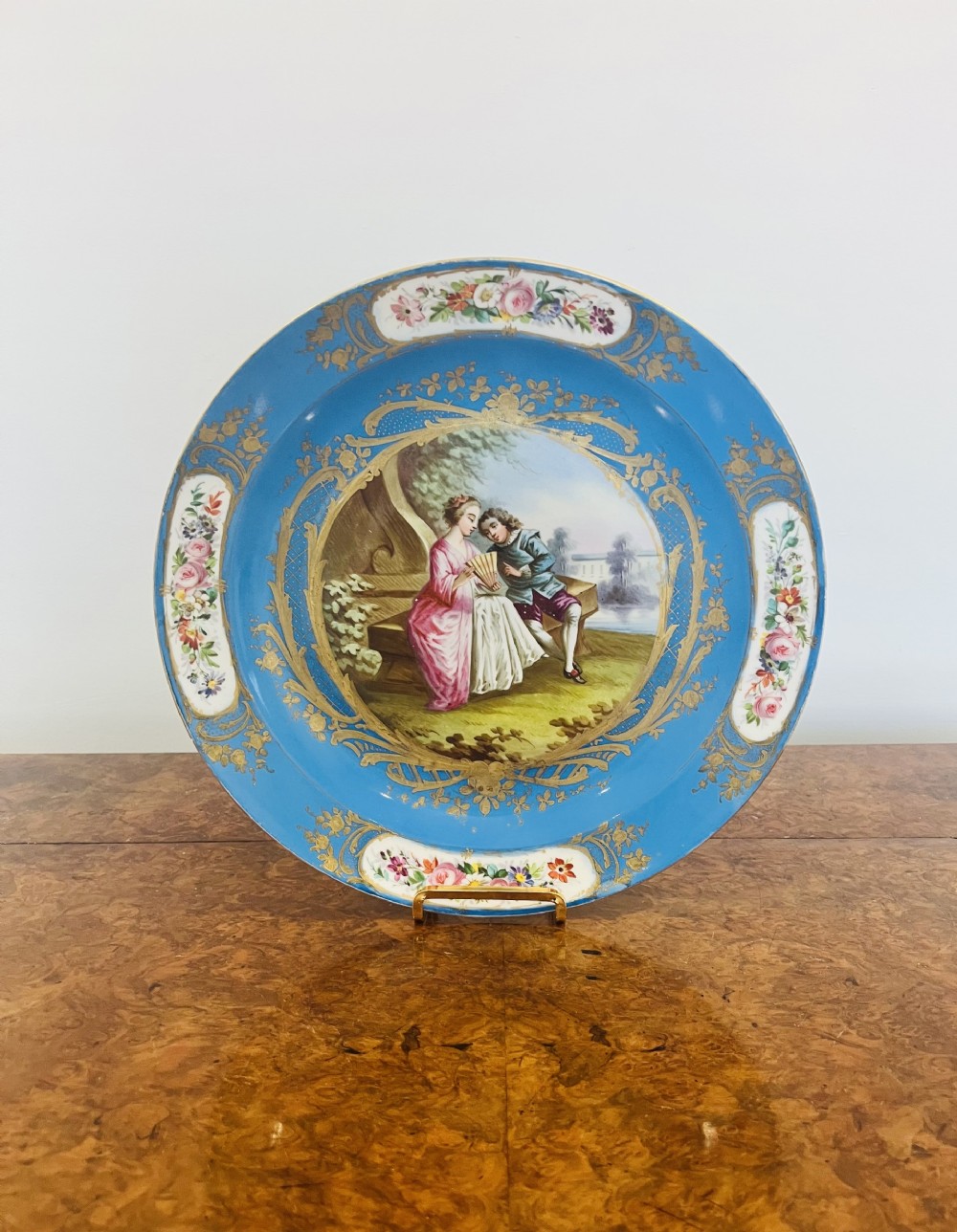 A circular sky blue porcelain dish with a hand-painted central vignette of a couple in historic attire framed by gold gilding and floral reserves.