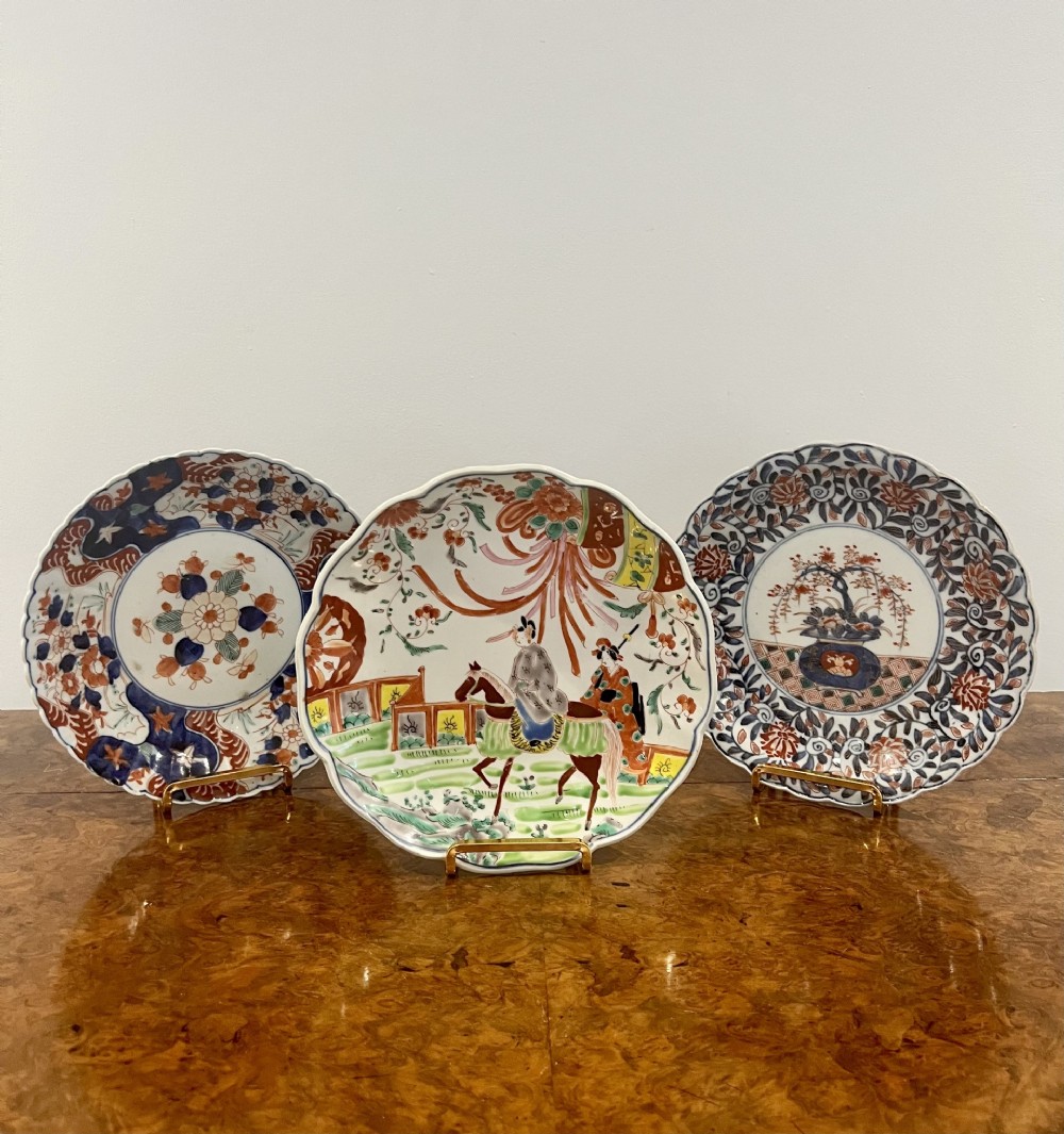 A set of three antique Japanese Imari porcelain dishes with scalloped rims, featuring hand-painted floral, figurative, and geometric motifs in blue, red, green, and gold.