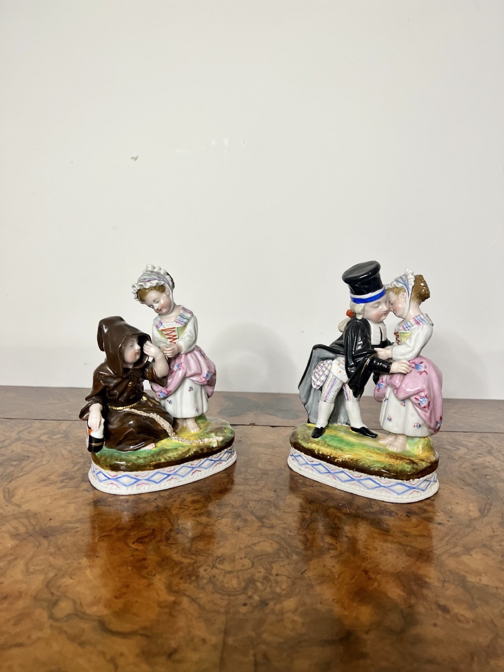 A pair of hand-painted antique continental porcelain figurines depicting romantic character figures standing on patterned oval bases.