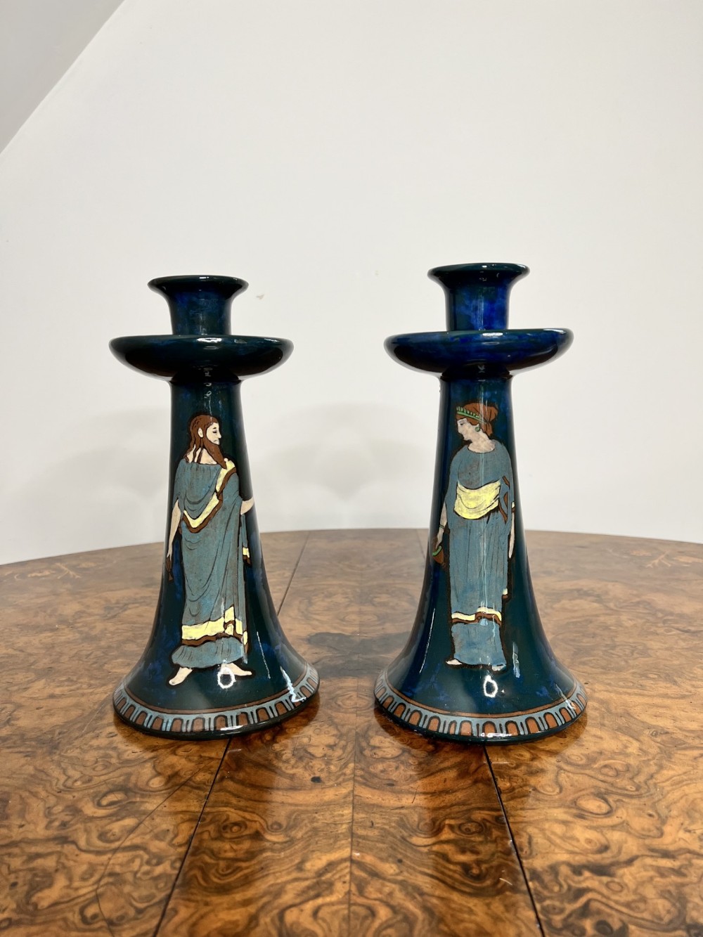A pair of flared dark blue glazed ceramic candlesticks decorated with hand-painted Classical figures and a patterned base border.
