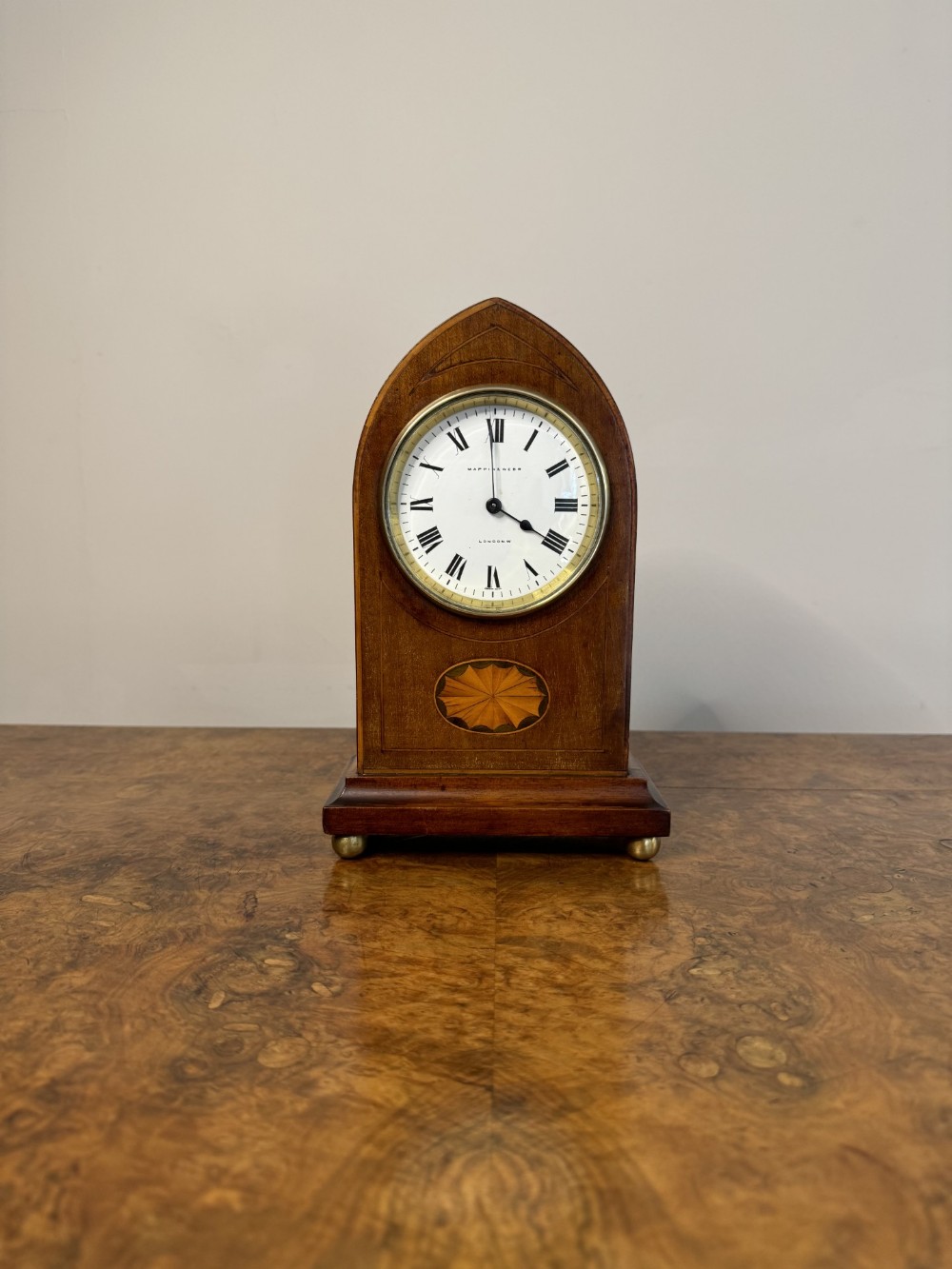 An Edwardian mahogany lancet mantel clock featuring satinwood oval inlay, a white enamel dial with Roman numerals, and brass ball feet.