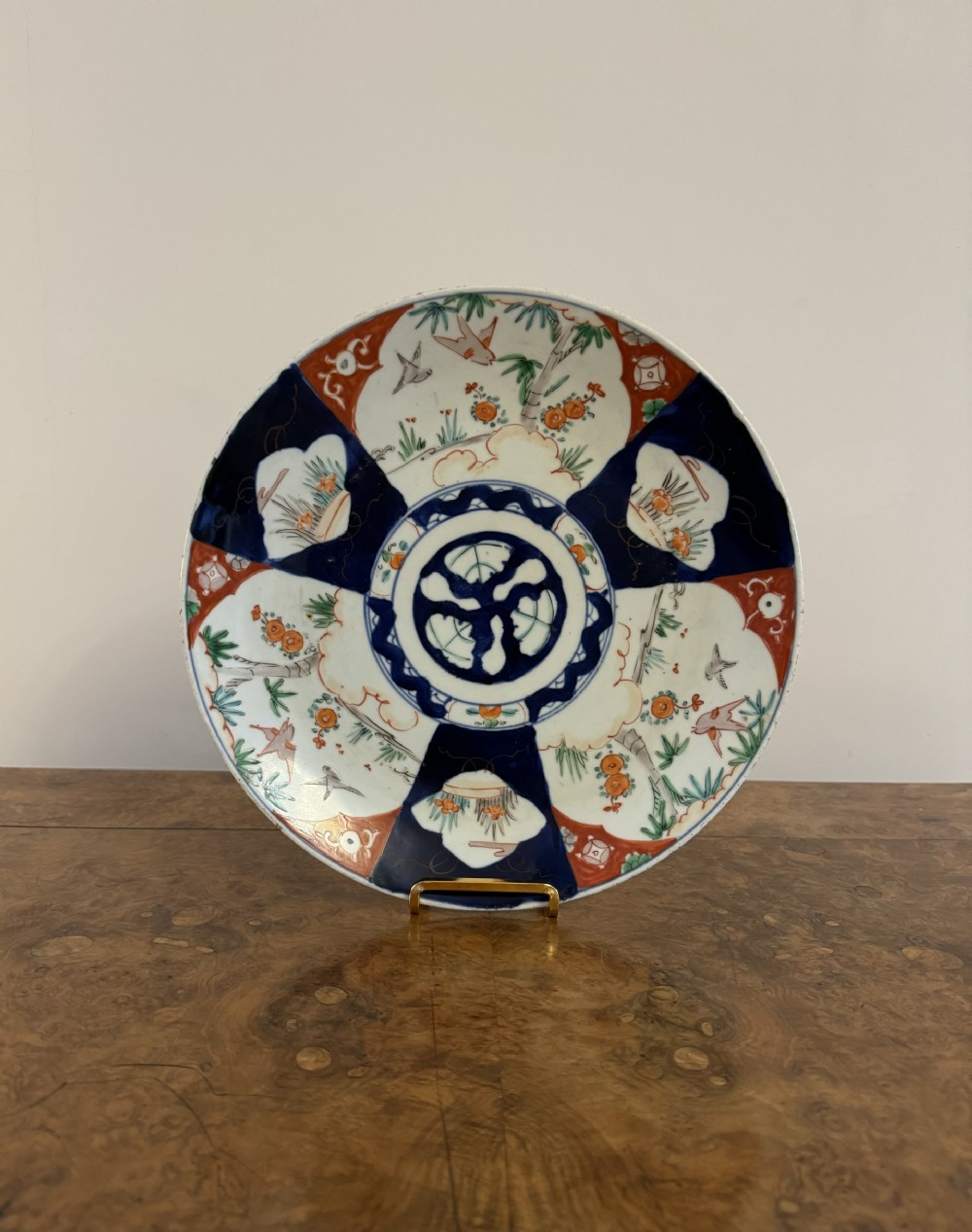 A circular antique Japanese Imari porcelain plate decorated with alternating navy blue, iron red, and white patterned panels featuring bird and botanical motifs around a central crest.