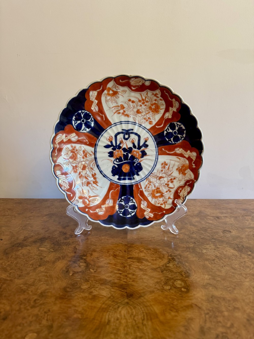 A circular Japanese Imari porcelain plate featuring a scalloped rim, hand-painted cobalt blue and iron-red floral motifs, and a central basket of flowers.