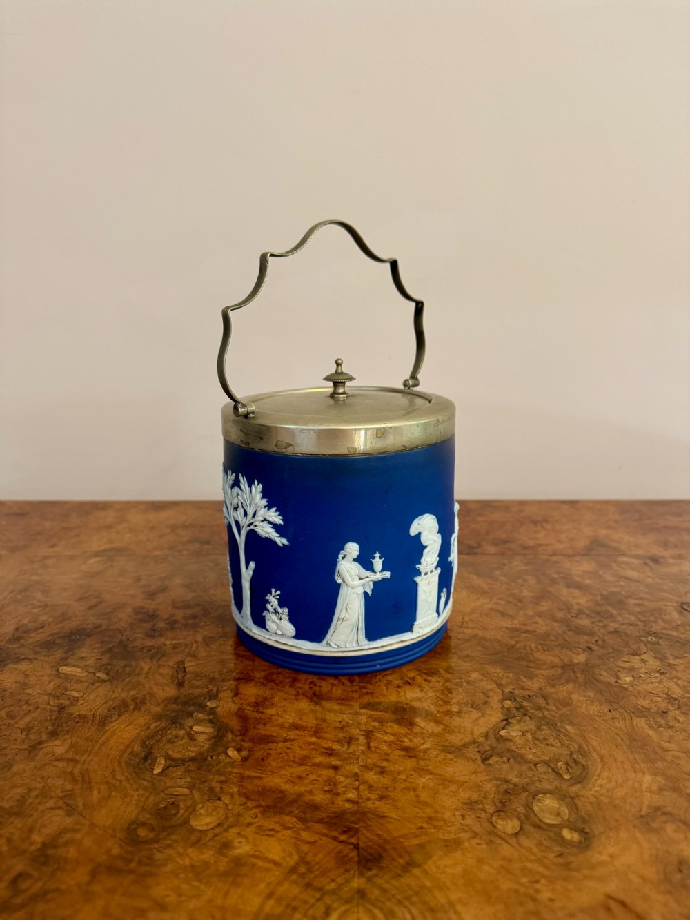 A cobalt blue Wedgwood Jasperware biscuit barrel with white classical bas-relief motifs, featuring a silver-plated rim, lid, and swing handle. View V