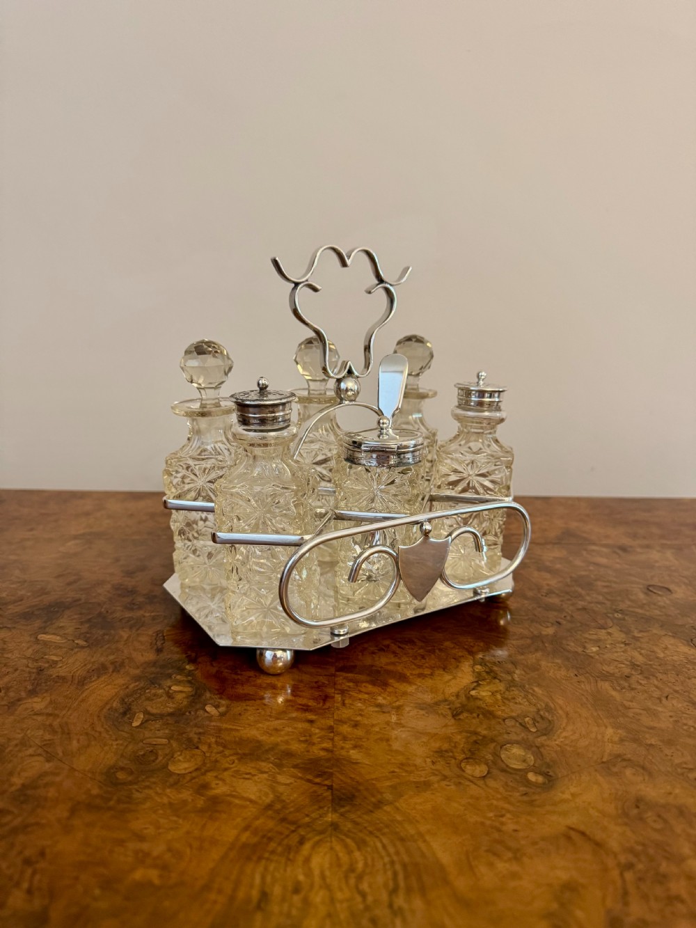 An Edwardian silver-plated frame holding six faceted cut-glass condiment bottles and stoppers on a polished wood surface.