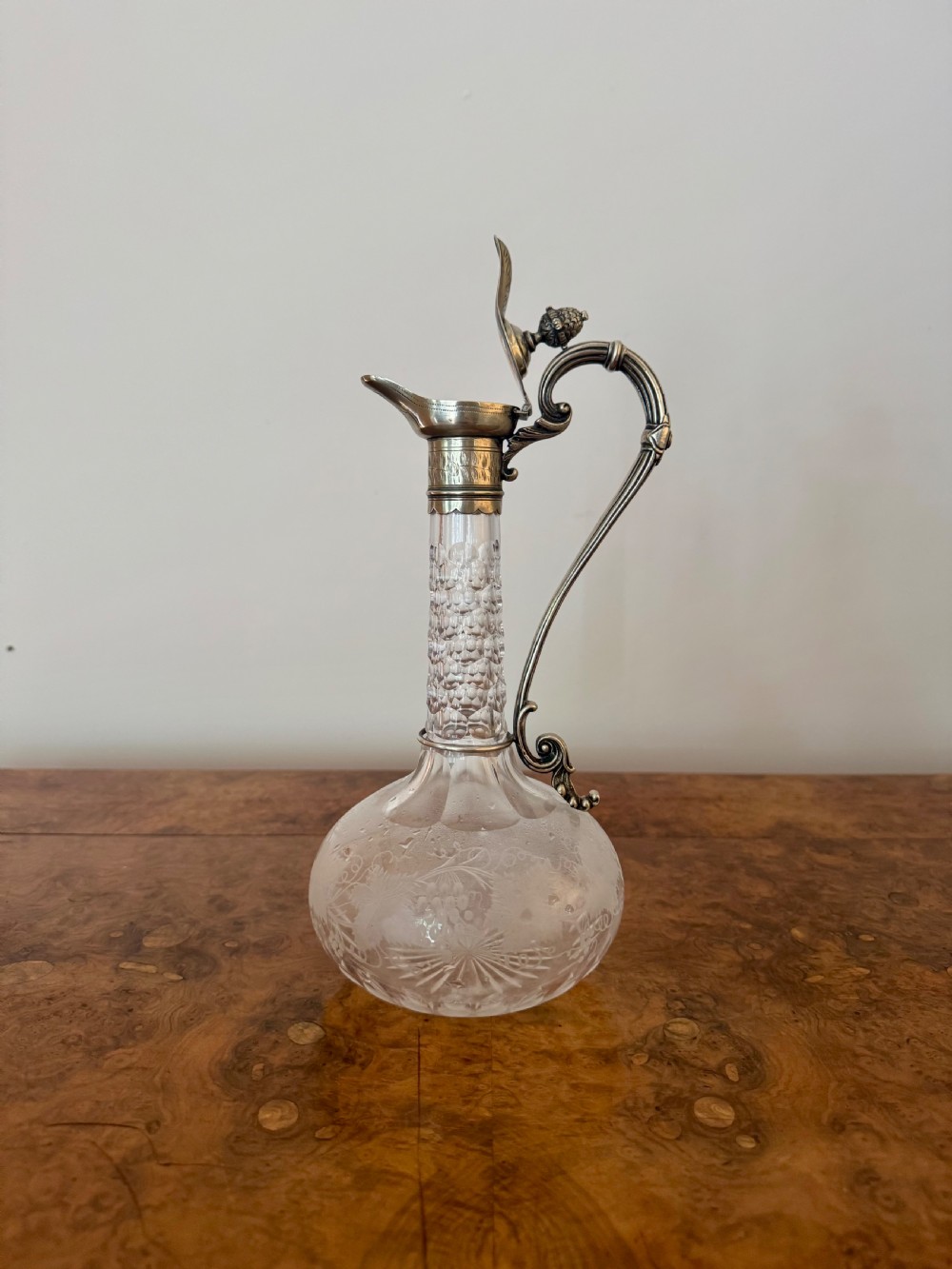 A Victorian clear etched glass claret jug featuring floral vine patterns, a textured glass neck, and a silver-plated collar with a scroll handle and finial lid. View V