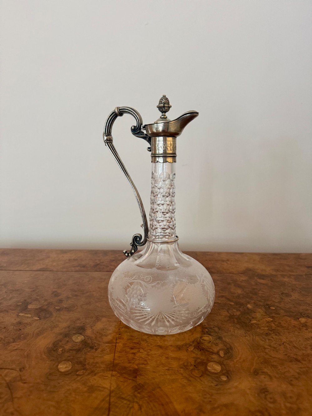 A Victorian clear etched glass claret jug featuring floral vine patterns, a textured glass neck, and a silver-plated collar with a scroll handle and finial lid. View VI