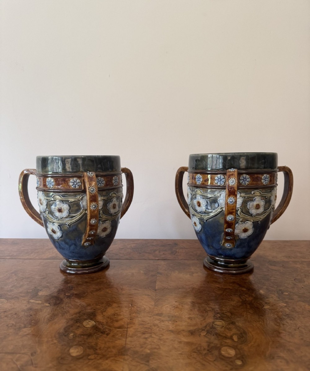 A pair of antique Royal Doulton glazed stoneware loving cups decorated with three handles and raised white floral reliefs against blue and brown tones.