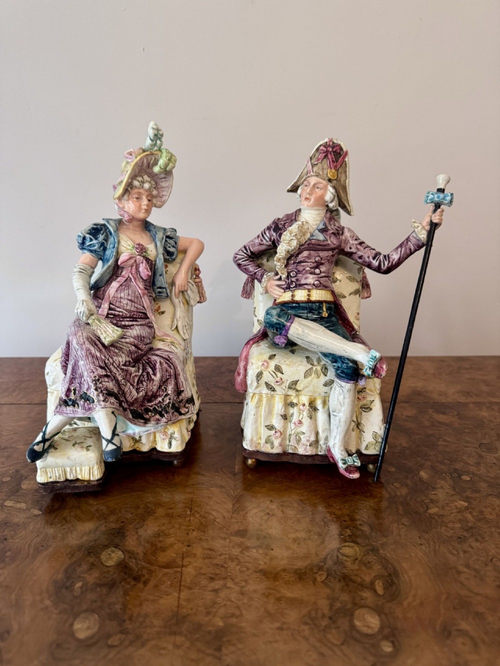 A pair of antique Victorian painted porcelain musical figures depicting a lady and a gentleman dressed in eighteenth-century costume seated on floral armchairs.