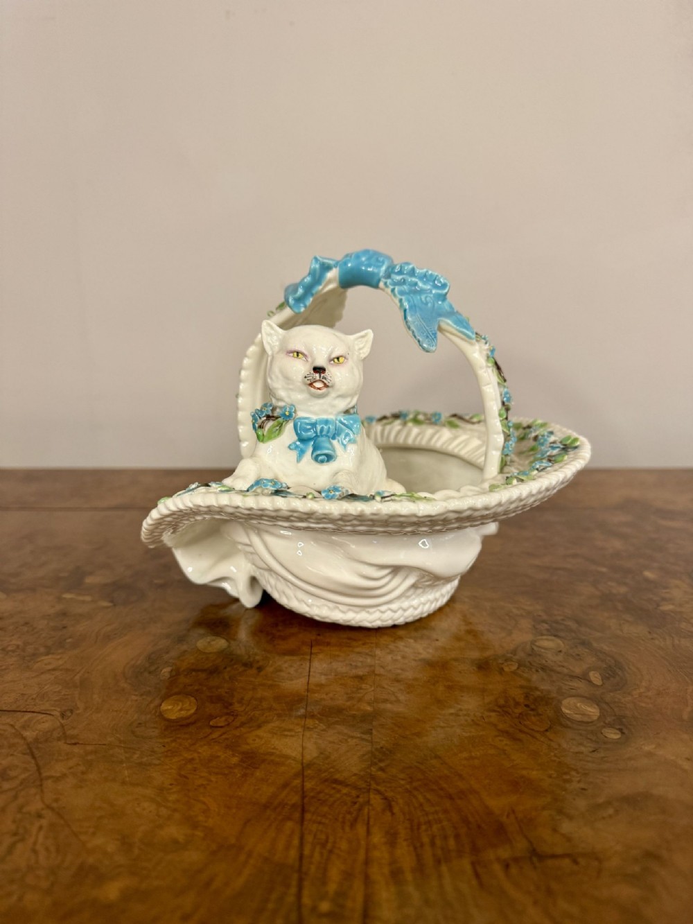 A Victorian white porcelain decorative basket featuring molded basketweave details, applied light blue ribbons, and a sculpted white cat figure seated inside.