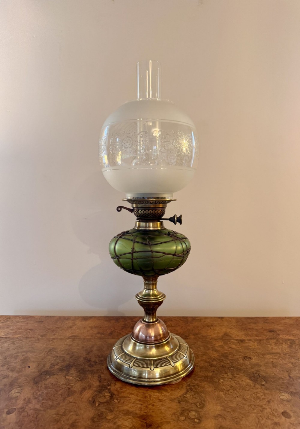 An antique Victorian oil lamp featuring a stepped brass and copper base, a green glass font wrapped in wirework, an etched frosted glass globe, and a clear glass chimney.