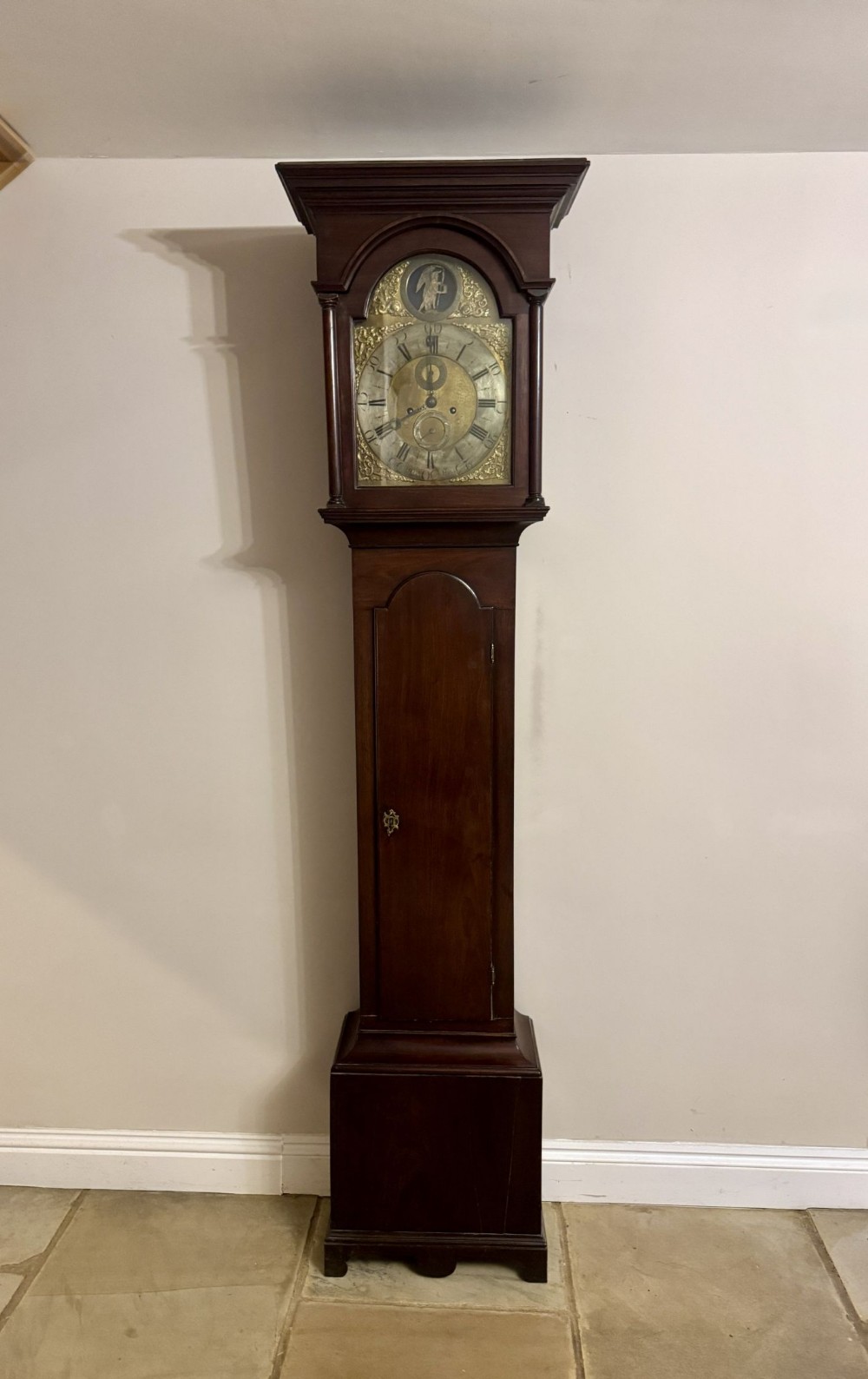 An antique George III longcase clock featuring a dark mahogany case, an arched glass hood with flanked columns, and an engraved brass dial.