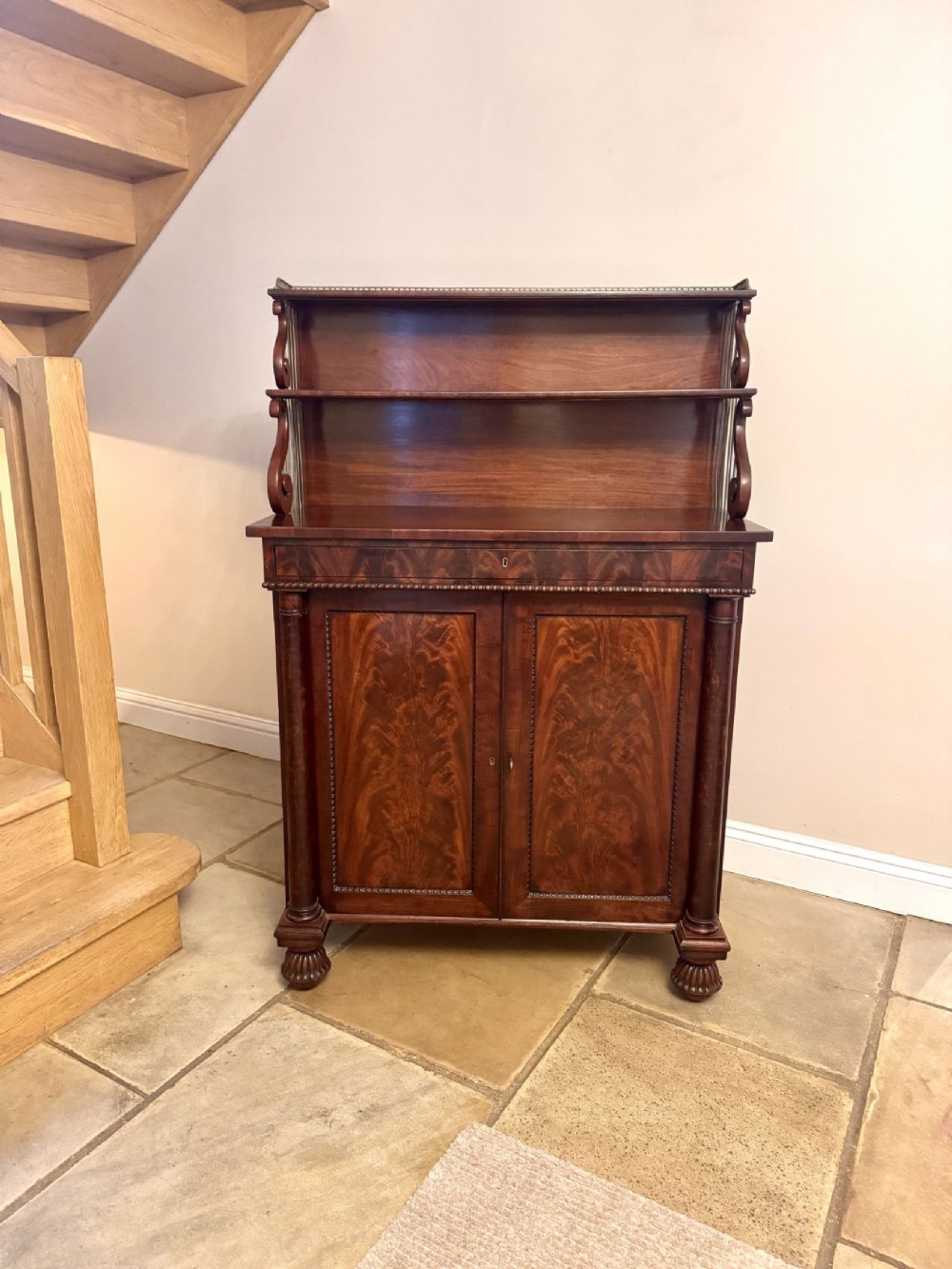 An antique Regency mahogany chiffonier cabinet featuring a raised open shelf, flame mahogany door panels, turned side columns, and bun feet.