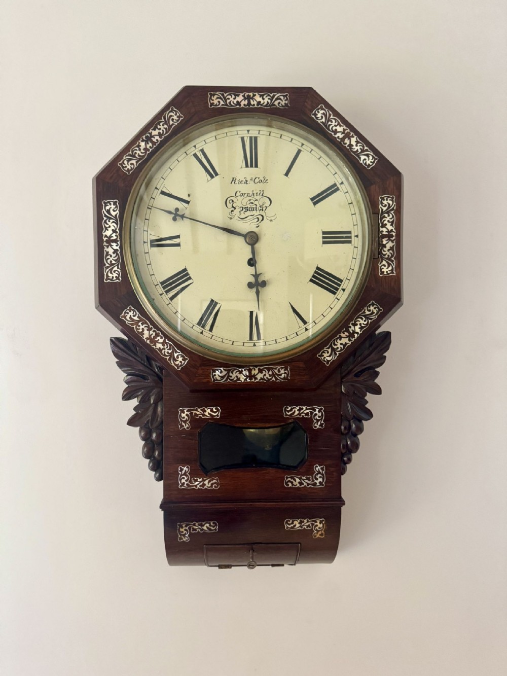 A Victorian octagonal rosewood drop dial wall clock featuring mother-of-pearl foliate inlay, carved side ear scrolls, and a glass pendulum viewing aperture.
