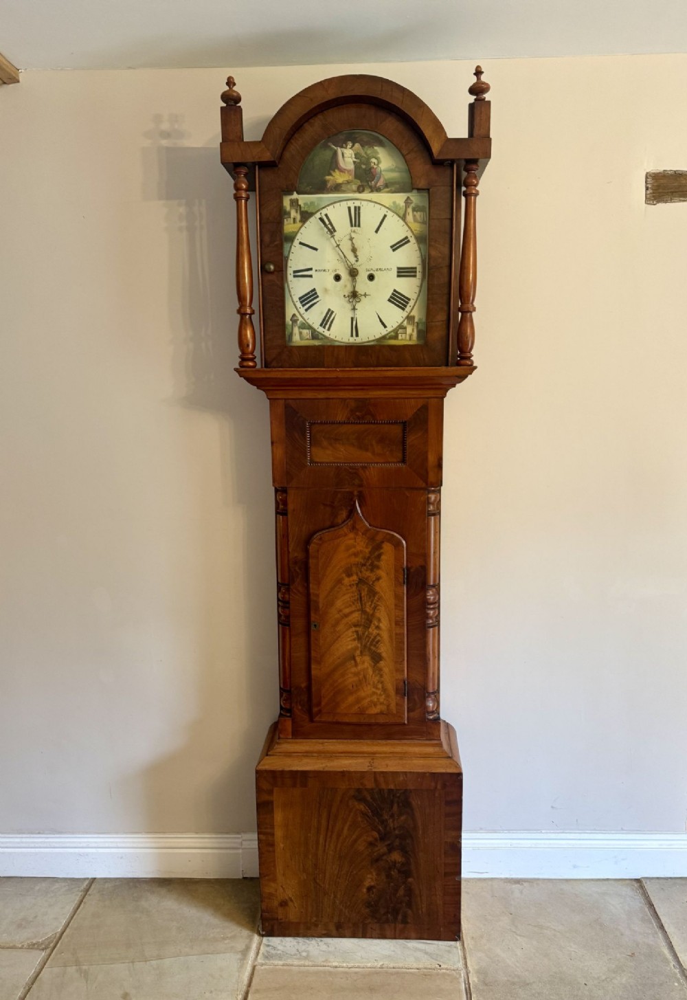 An antique 19th-century mahogany longcase clock featuring a painted arch dial, turned column accents, finials, and a flame mahogany veneer door.