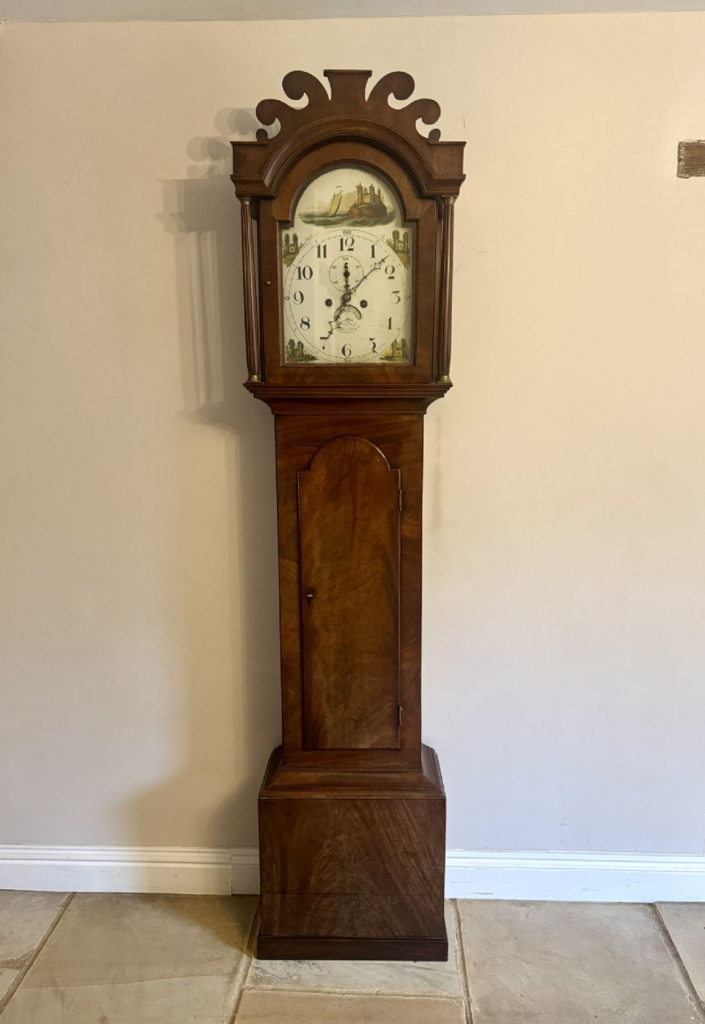 A George III mahogany longcase clock featuring a carved scroll pediment hood, painted arch dial with nautical illustration, flame mahogany trunk door, and plinth base.