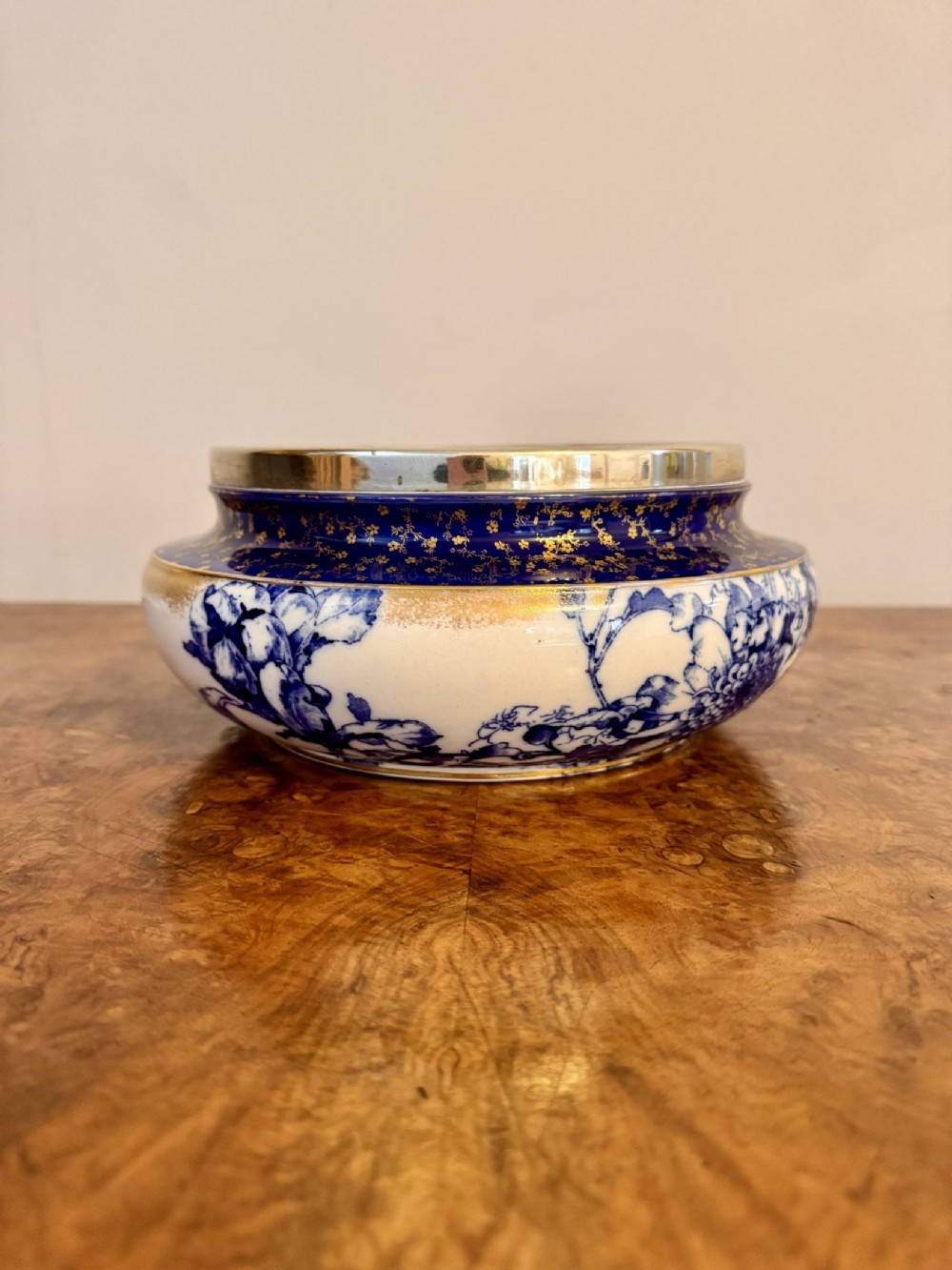 A rounded ceramic bowl featuring blue and white floral patterns, a deep cobalt blue shoulder highlighted with gold accents, and a polished silver rim.
