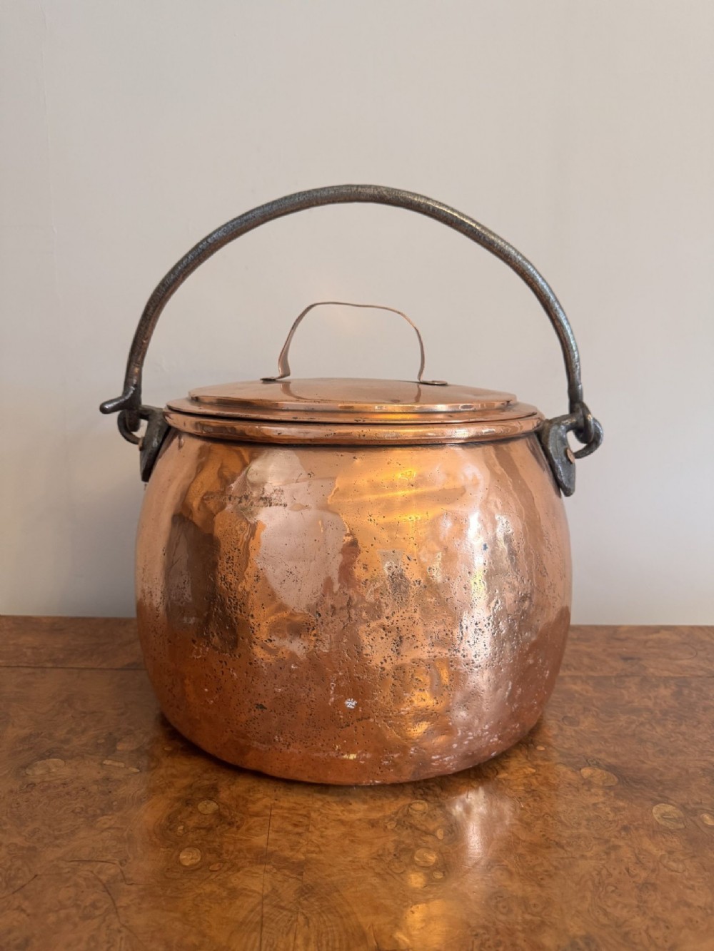 A large round hand-hammered copper pot with a fitted copper lid and a dark iron swing handle.