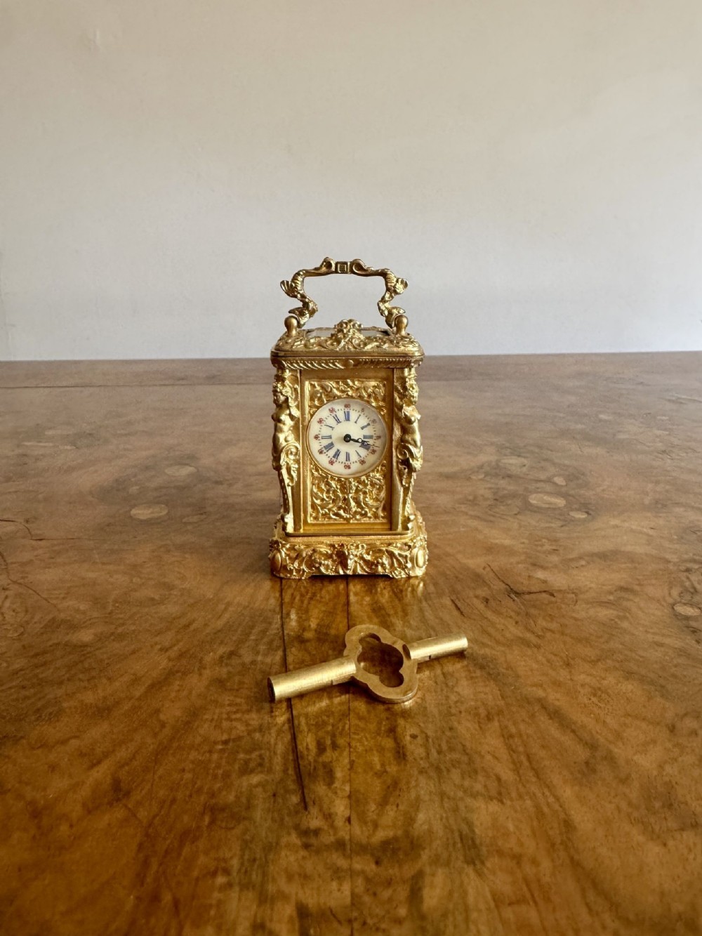 An ornate gilded brass miniature carriage clock featuring detailed castings, an enamel dial, and a matching brass winding key.