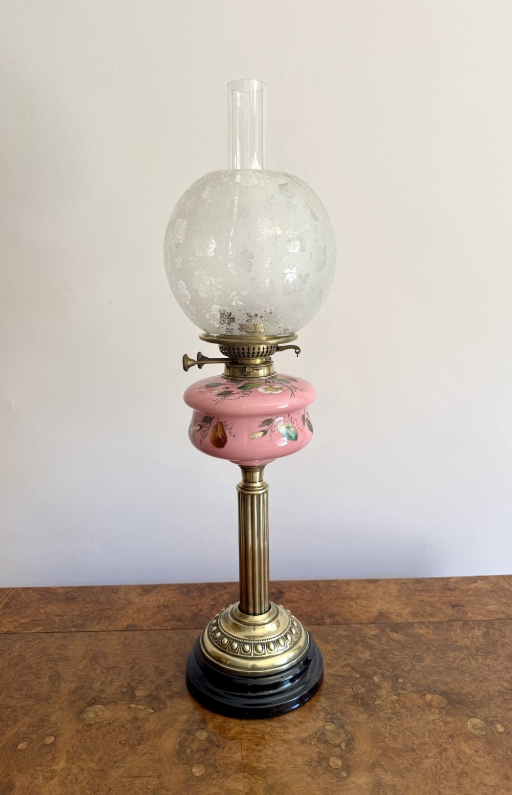 A Victorian oil lamp with a black ceramic base, fluted brass column, hand-painted pink glass reservoir, frosted globe shade, and clear chimney.