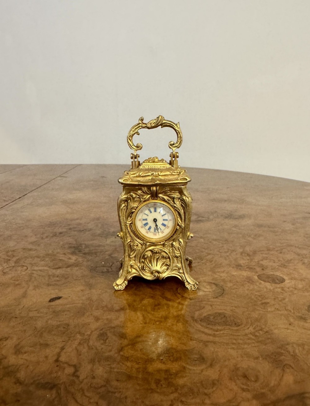 A miniature Victorian gilded brass carriage clock featuring Rococo foliage carvings, an ornate hinged top handle, and a circular white enamelled dial with Roman numerals.