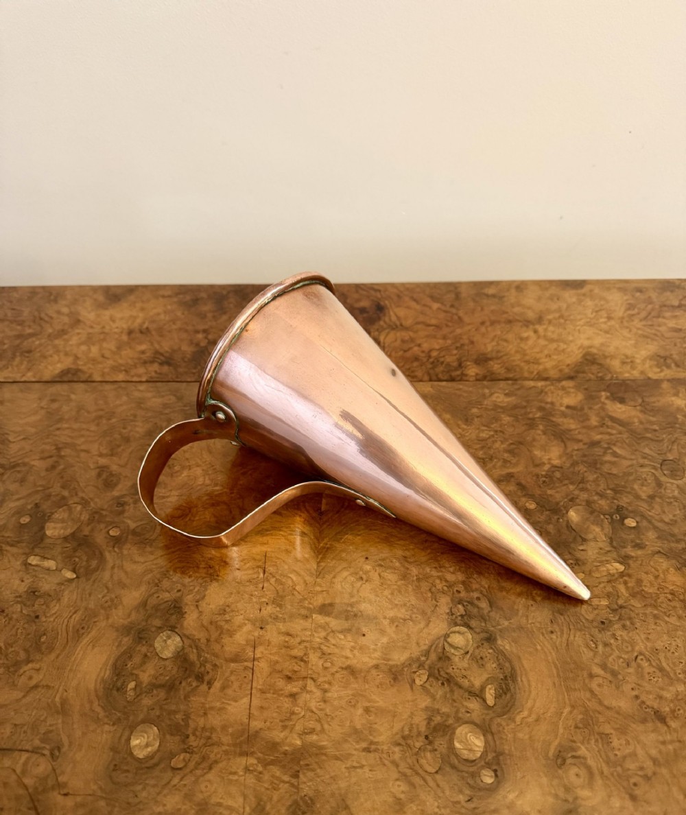 A conical antique George III copper measure with a curved strap handle resting horizontally on a polished wood surface.