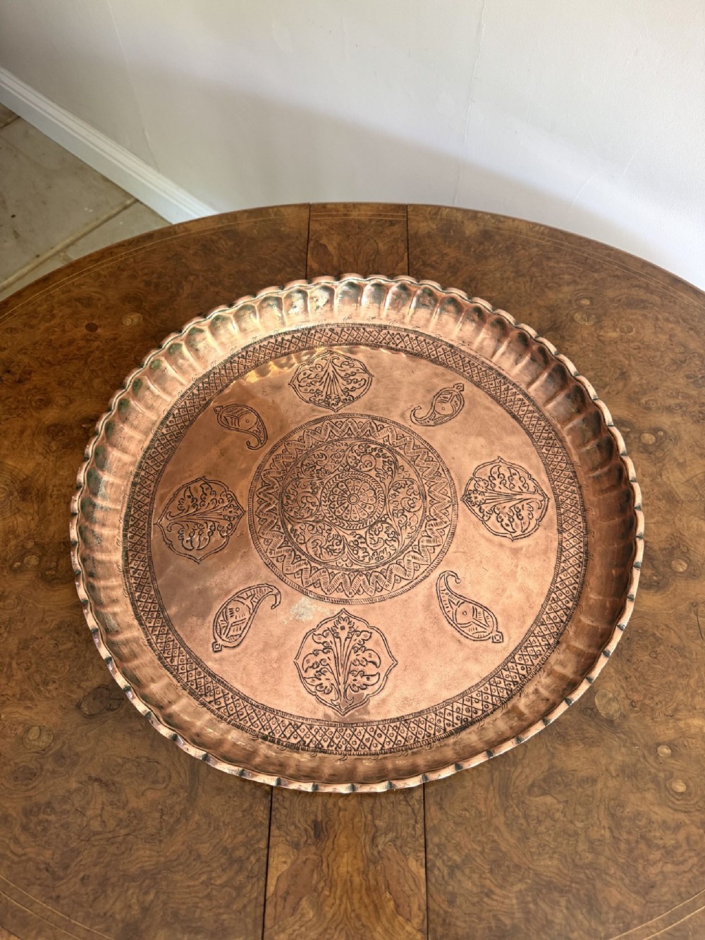 A large circular copper tray with a scalloped fluted rim and detailed hand-engraved floral, paisley, and geometric patterns across the surface.