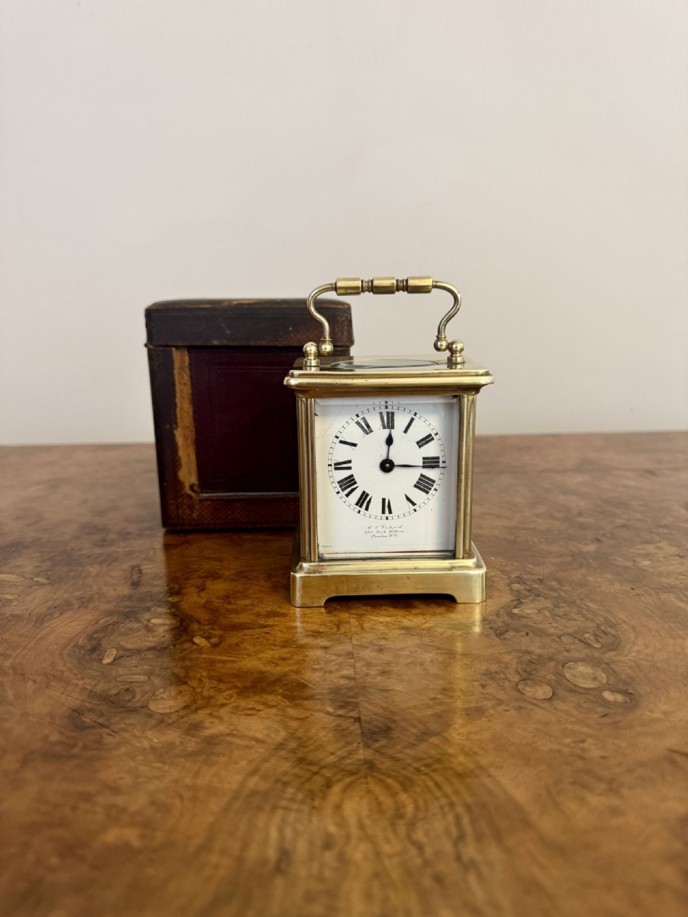An antique Victorian brass carriage clock featuring a white enamel dial with Roman numerals, glass side panels, and a top carrying handle alongside its brown leather transport case.