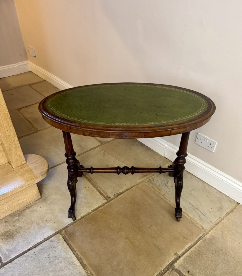 An oval Victorian walnut writing table featuring a green leather inset top with gold tooling, turned end supports, a turned stretcher, and carved legs.