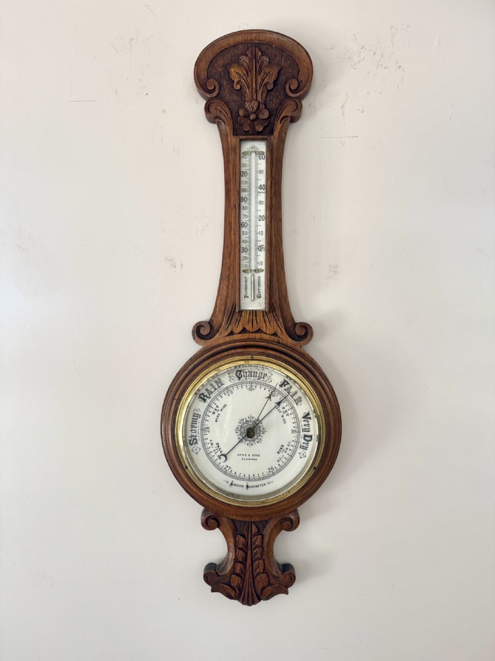 An Edwardian carved oak aneroid barometer featuring an upper thermometer tube and lower circular dial with brass bezel.
