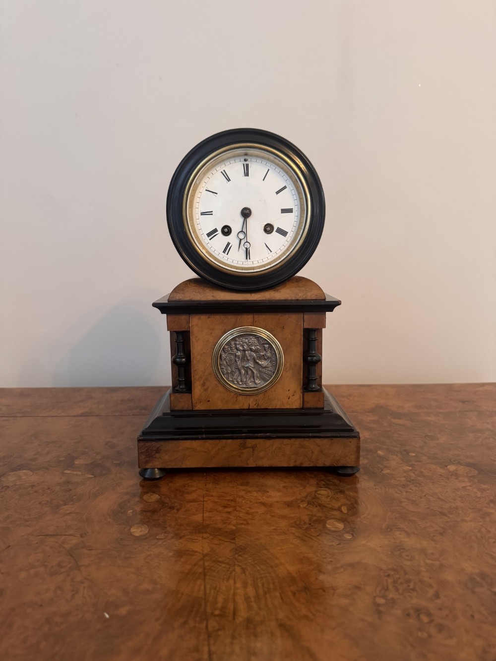 A Victorian burr walnut mantel clock featuring an upper circular white enamel dial with Roman numerals, ebonised side columns, and a central circular relief medallion inset into the pedestal base.