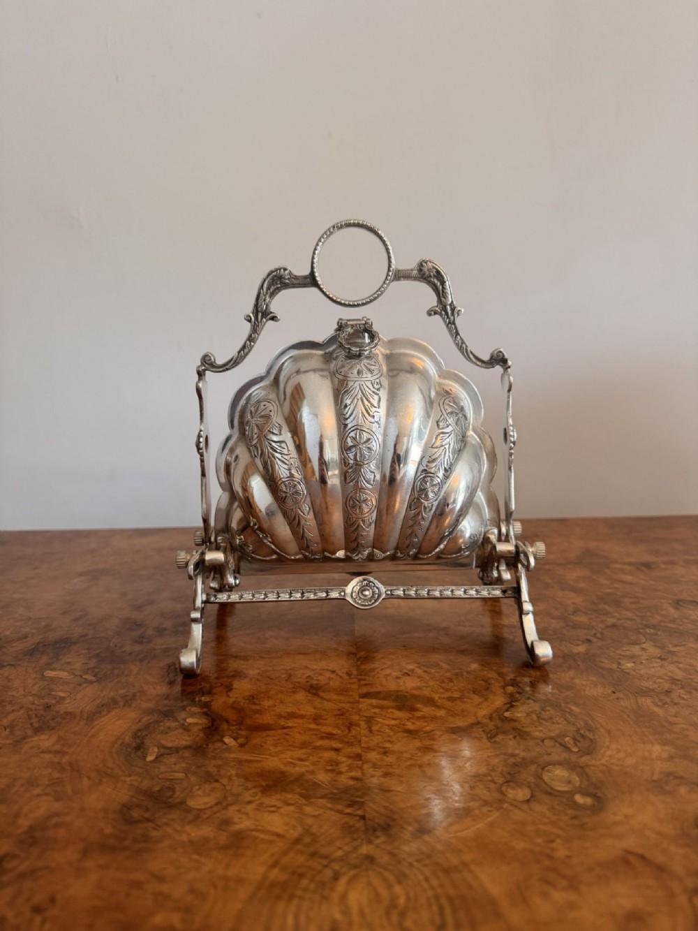 A silver-plated shell-shaped folding biscuit box mounted on a decorative cast metal frame with a ring top handle.