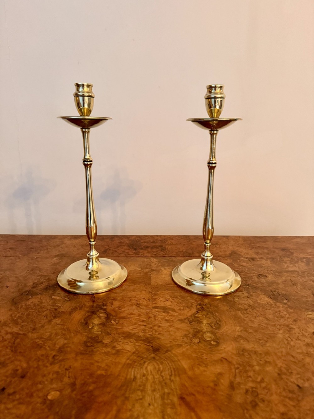 A pair of slender antique brass candlesticks with turned stems, circular drip pans, and circular bases resting on a polished wooden surface.
