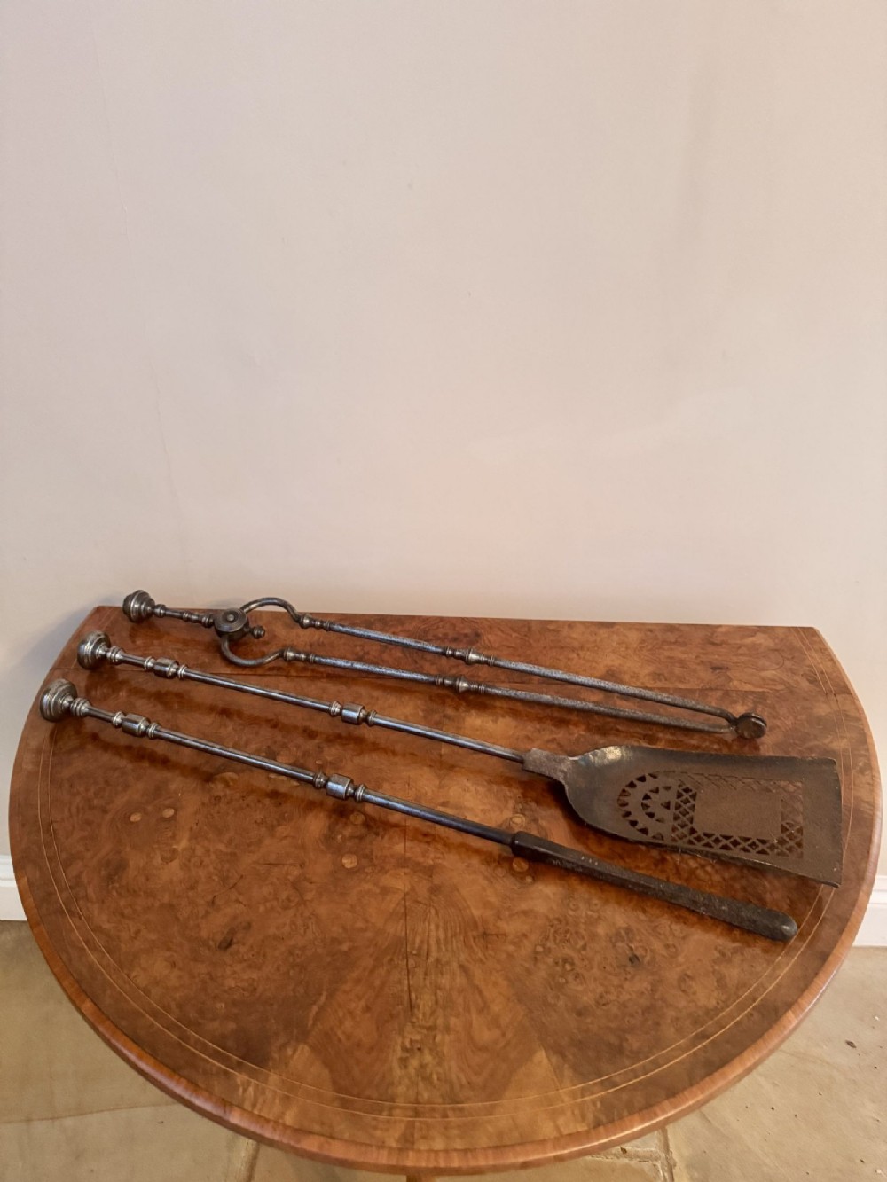 A three-piece set of nineteenth-century steel fire irons including a poker, tongs, and a shovel with a pierced blade resting on a wooden surface.