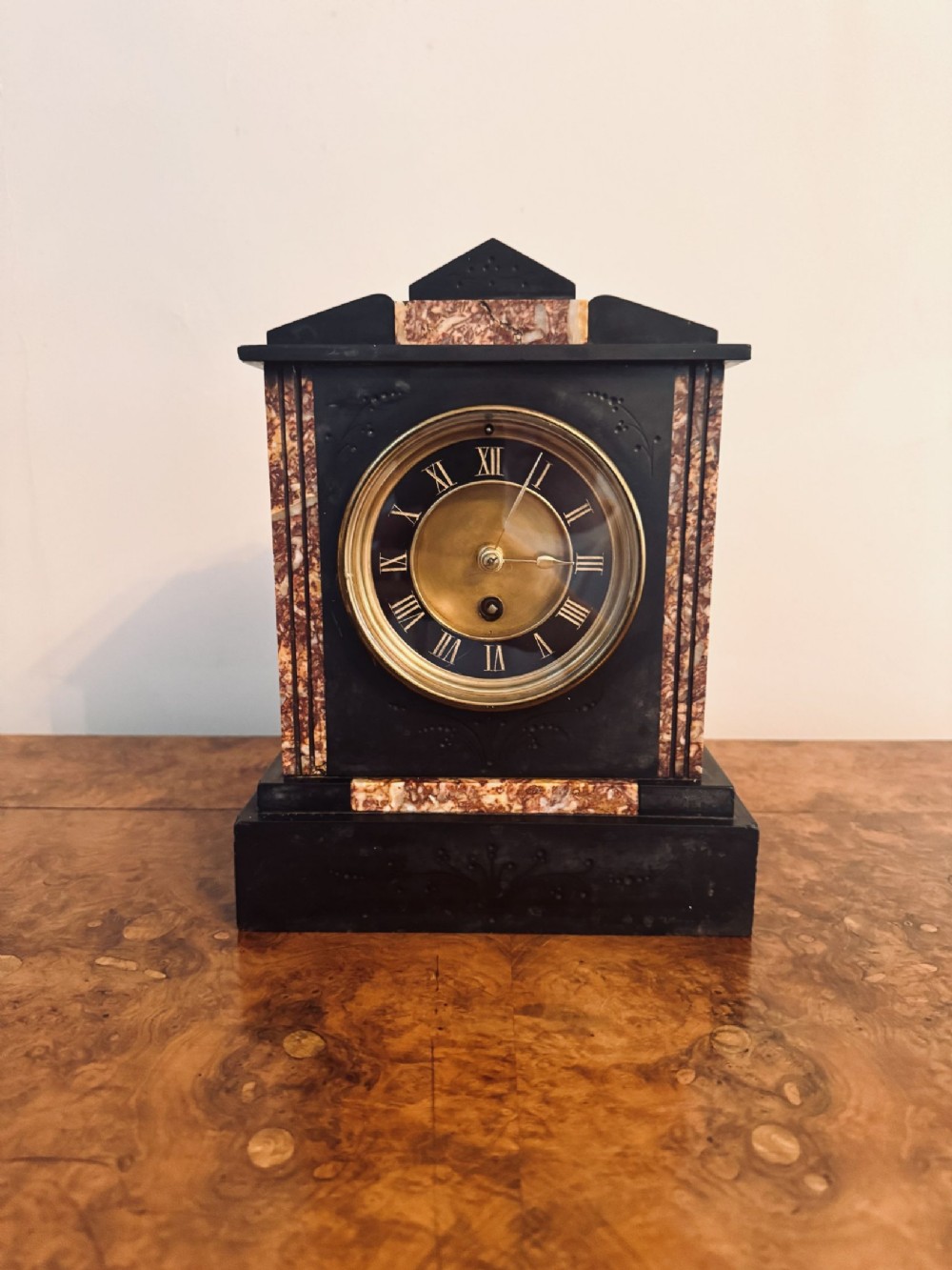 A Victorian black slate mantel clock featuring reddish-brown marble inlays, a gabled pediment top, and a circular brass dial with Roman numerals.