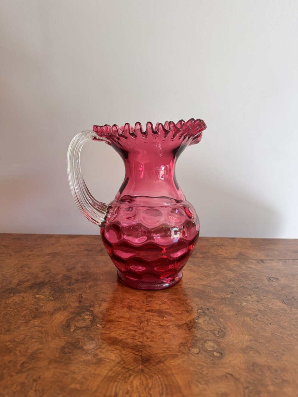 A Victorian cranberry glass jug featuring a crimped frilled rim, an optic dimpled body, and a clear ribbed glass handle.