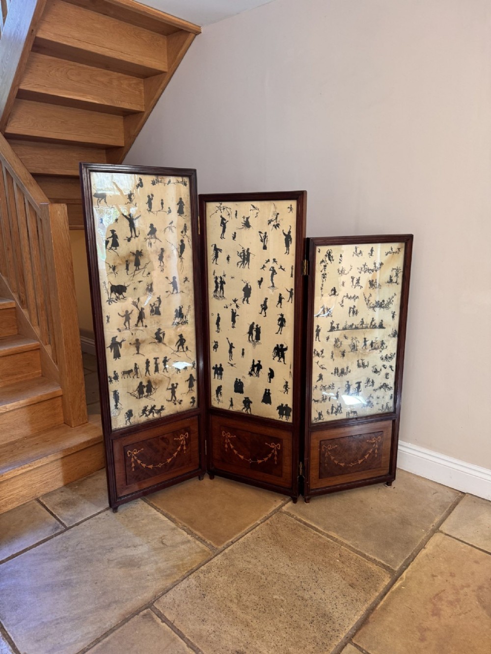 A three-panel Victorian mahogany folding draft screen with graduated heights, marquetry swags at the base, and printed silhouette illustrations.