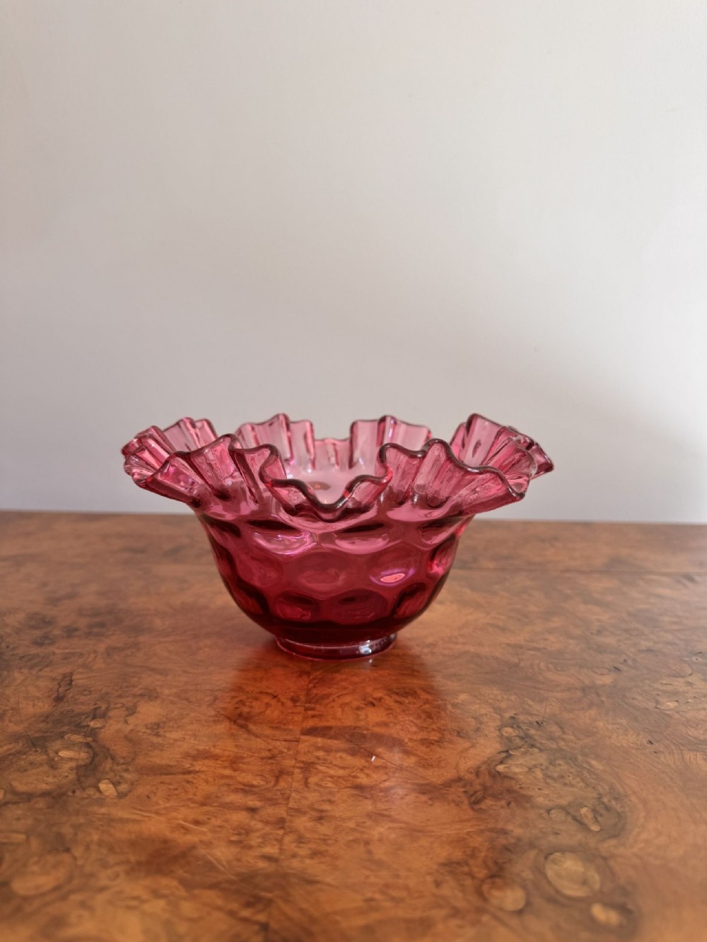 A small antique cranberry glass bowl featuring a crimped ruffled rim and a textured thumbprint pattern.