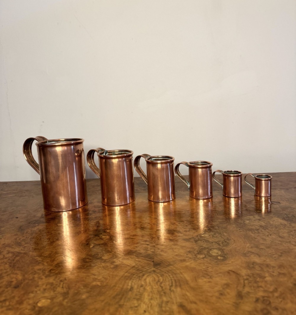 A graduated set of six polished Victorian copper liquid measures with strap handles, arranged in descending size from left to right on a wooden surface.