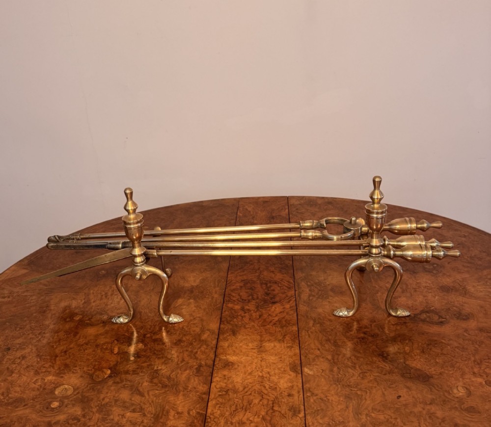 A set of three polished brass fire irons resting horizontally across a pair of matching brass fire dogs with urn-shaped finials.